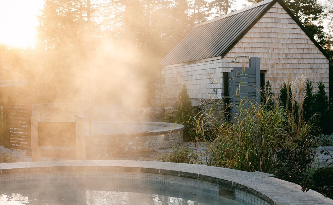 A tranquil hot tub at Oceanstone Resort and Spa, mist rising amid stones, lush plants, and sunlit trees beside a charming wooden shed.