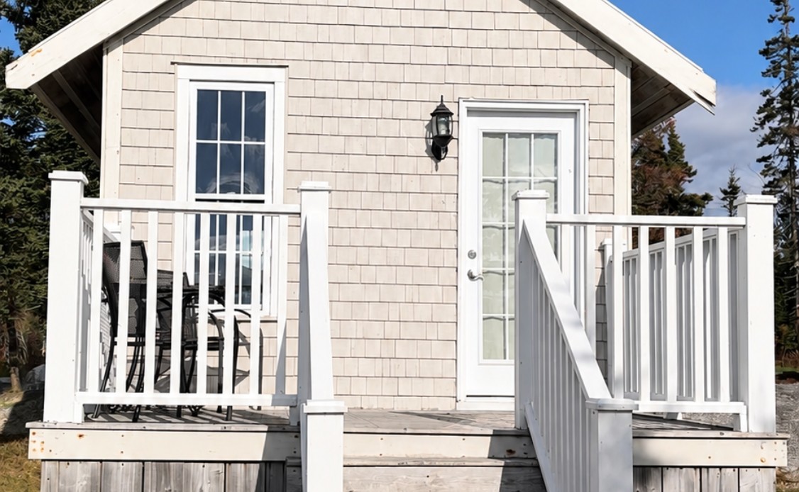 15 A cozy beige cottage with a white porch and outdoor chairs, nestled amid grass and trees for a serene, relaxing resort escape.