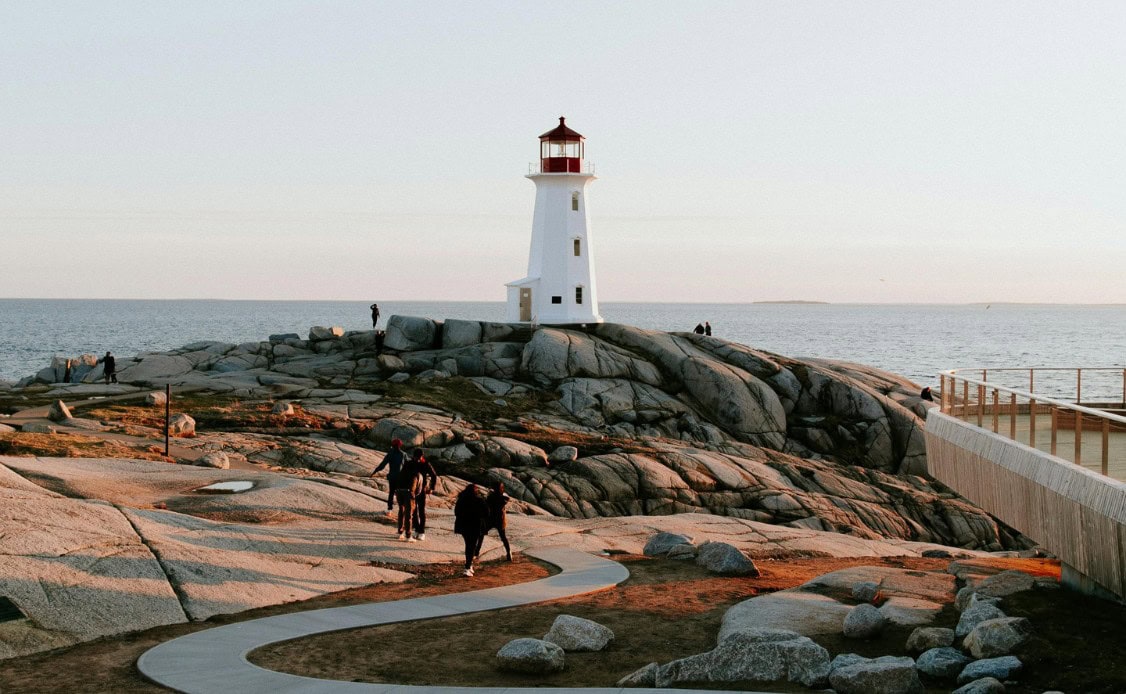 A peaceful white lighthouse with a red top overlooks the ocean as visitors enjoy sunlit paths and tranquil rocky shores under clear skies.