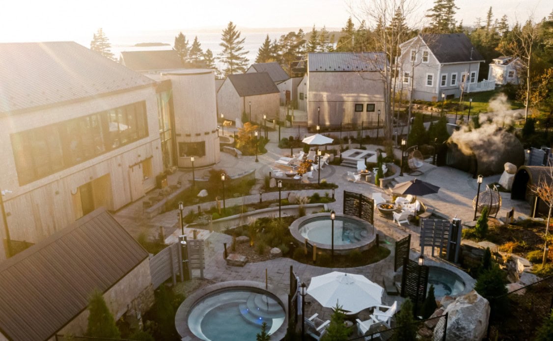 A tranquil spa resort with round hot tubs, cozy loungers, gardens, and steam rising, nestled by forest and water for total relaxation.