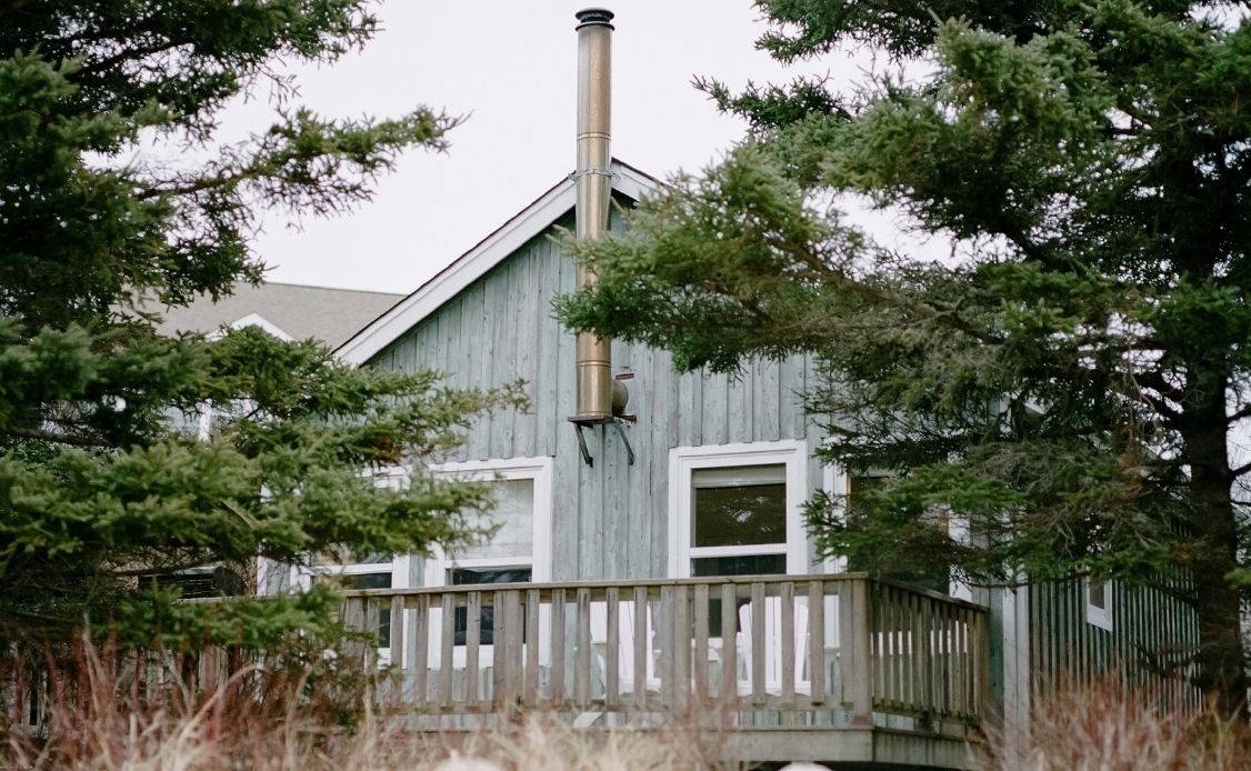 1 A peaceful, light-gray wooden cabin with a charming balcony and metal chimney nestles quietly amid tall evergreen trees, inviting relaxation. Two large front-facing windows offer scenic views of the tranquil surroundings. In the foreground, soft dried grass and twigs add to the natural ambiance, while an overcast sky above enhances the serene atmosphere of Oceanstone Resort and Spa.