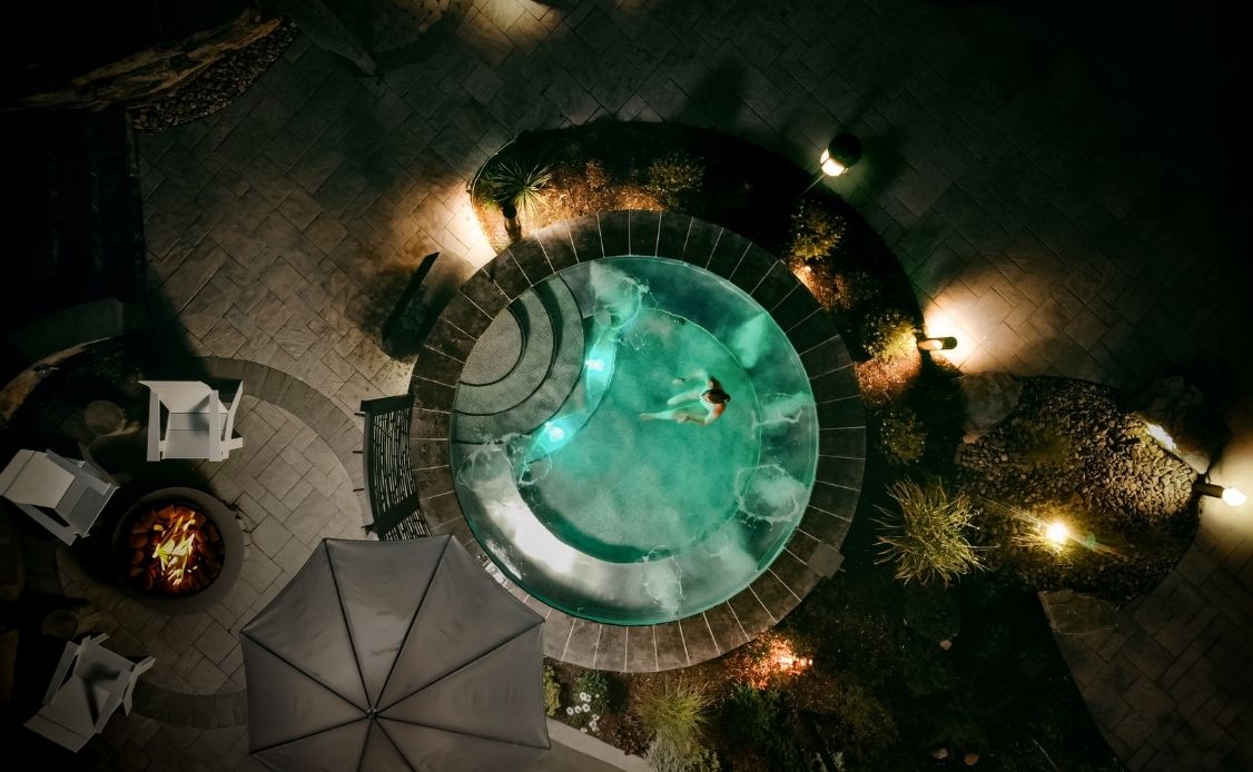 2 A tranquil aerial view at night captures a circular hot tub with a single guest unwinding, illuminated by gentle poolside lamps. Surrounded by elegant stone tiles, lush plants, cozy patio furniture, a glowing fire pit, and the shelter of a large umbrella, the scene reflects the peaceful and rejuvenating atmosphere of Oceanstone Resort and Spa.