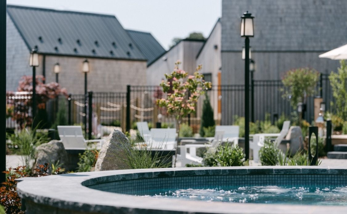 5 A bubbling round hot tub sits invitingly in the foreground, nestled among lush greenery and comfortable lounge chairs, with gentle lamp posts adding a soft glow. A sleek black metal fence frames the tranquil space. Behind, modern gray buildings with dark roofs blend seamlessly into the peaceful setting, all beneath a clear and calming sky at Oceanstone Resort and Spa.