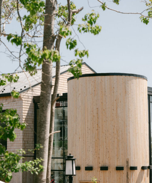 A tranquil modern building at Oceanstone Resort and Spa features a tall, light wood cylindrical structure and expansive glass windows, topped with a shingled roof. The peaceful setting is enhanced by leafy tree branches gently framing the view under a clear sky. A black outdoor lantern rests quietly at the bottom center, completing the serene atmosphere.