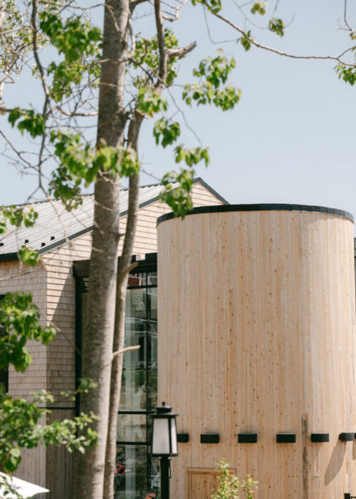 A tranquil modern building at Oceanstone Resort and Spa features a tall, light wood cylindrical structure and expansive glass windows, topped with a shingled roof. The peaceful setting is enhanced by leafy tree branches gently framing the view under a clear sky. A black outdoor lantern rests quietly at the bottom center, completing the serene atmosphere.